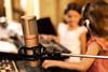 Two young girls wear headphones as they sit in front of a microphone and sound board