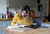 A woman writes in a notepad as she sits at her kitchen table in front of a laptop