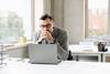 A male office worker clasps his hands together as he reads something on his laptop