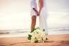 A newly-married couple hold hands on a beach as the bride's bouquet lies on the sand