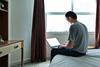 A man sits on the edge of a bed typing on a laptop