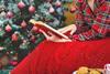 A woman wearing festive pyjamas and a blanket reads a book by a Christmas tree