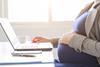 A pregnant woman rests her hand on her baby bump as she works at a laptop