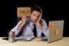 Exasperated man holding up a sign reading 'Help' as he sits in front of a laptop