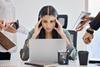 A woman holds her head in her hands feeling overwhelmed as she looks at her laptop, while colleagues try to get her attention