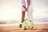A newly-married couple hold hands on a beach as the bride's bouquet lies on the sand