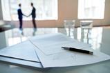 A signed contract sits on a boardroom table as businessmen shake hands in the background