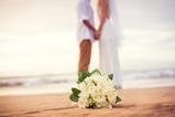 A newly-married couple hold hands on a beach as the bride's bouquet lies on the sand