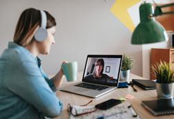 A woman wearing headphones and holding a drink talks to her colleague on a video call