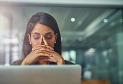 A young woman sits in front of her laptop with her eyes closed and her hands over her face