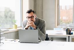 A male office worker clasps his hands together as he reads something on his laptop