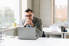 A male office worker clasps his hands together as he reads something on his laptop