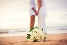 A newly-married couple hold hands on a beach as the bride's bouquet lies on the sand
