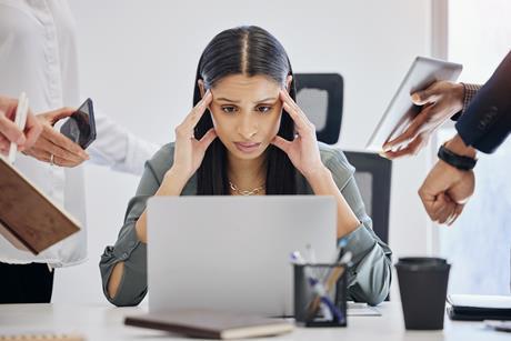 A woman holds her head in her hands feeling overwhelmed as she looks at her laptop, while colleagues try to get her attention