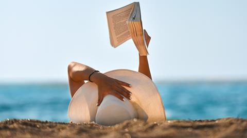 A woman lies on the sand wearing a sun hat reading a book