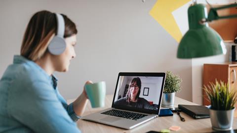 A woman wearing headphones and holding a drink talks to her colleague on a video call