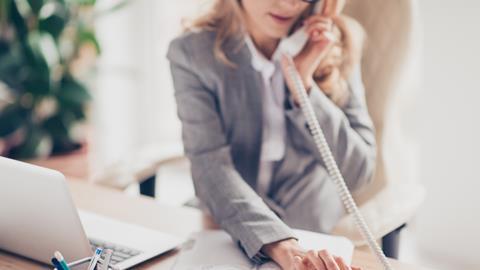 An anonymous woman in workwear dials a number on her desk phone as she works at her laptop