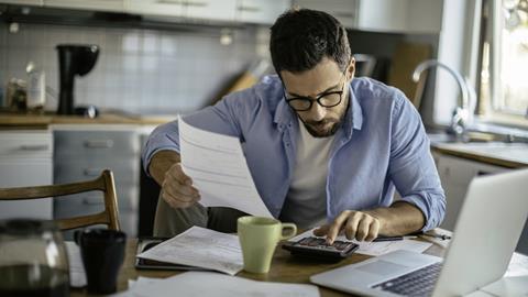 A man works through household bills using a calculator at his kitchen table