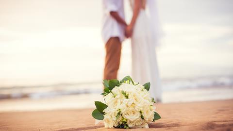 A newly-married couple hold hands on a beach as the bride's bouquet lies on the sand