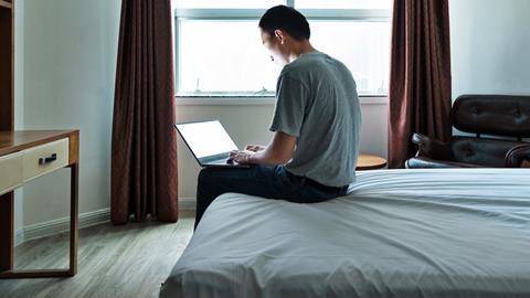 A man sits on the edge of a bed typing on a laptop