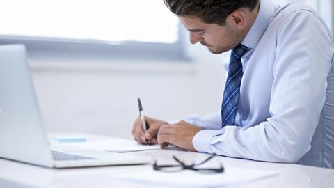 Man signing document sat at desk in front of laptop