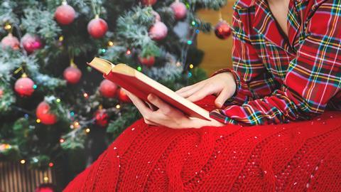 A woman wearing festive pyjamas and a blanket reads a book by a Christmas tree