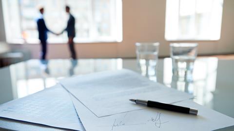 A signed contract sits on a boardroom table as businessmen shake hands in the background