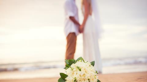 A newly-married couple hold hands on a beach as the bride's bouquet lies on the sand