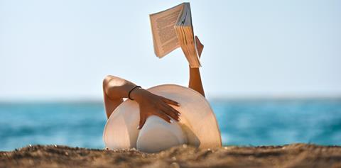 A woman lies on the sand wearing a sun hat reading a book