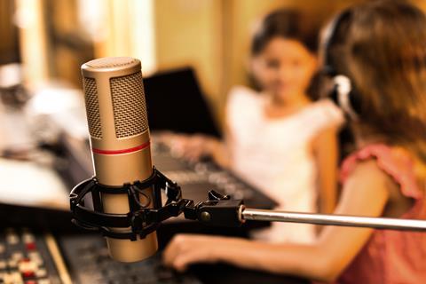 Two young girls wear headphones as they sit in front of a microphone and sound board