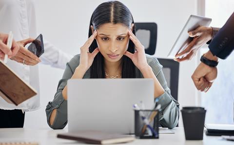 A woman holds her head in her hands feeling overwhelmed as she looks at her laptop, while colleagues try to get her attention
