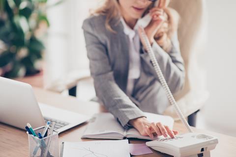 An anonymous woman in workwear dials a number on her desk phone as she works at her laptop