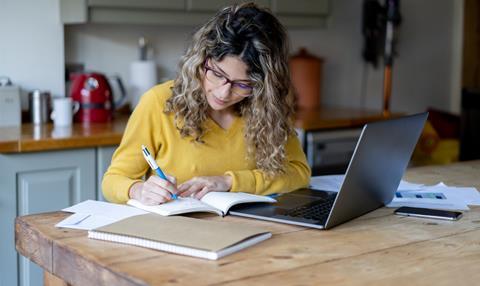 A woman writes in a notepad as she sits at her kitchen table in front of a laptop