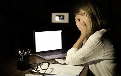 A young woman sits with her head in her hands in front of a laptop in a dark office