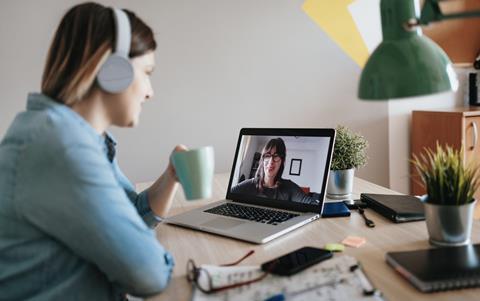A woman wearing headphones and holding a drink talks to her colleague on a video call