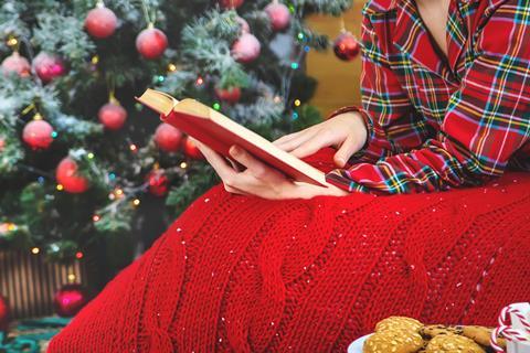 A woman wearing festive pyjamas and a blanket reads a book by a Christmas tree