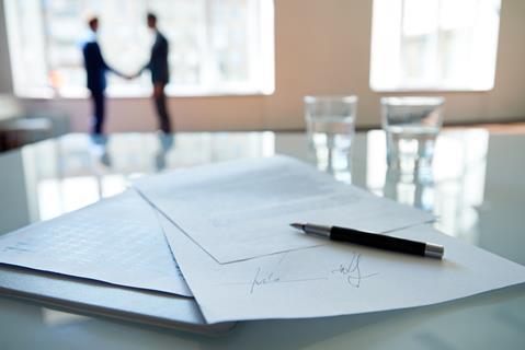 A signed contract sits on a boardroom table as businessmen shake hands in the background