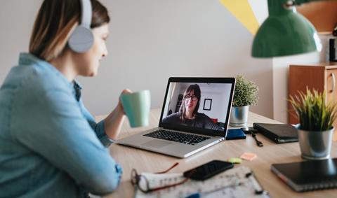 A woman wearing headphones and holding a drink talks to her colleague on a video call