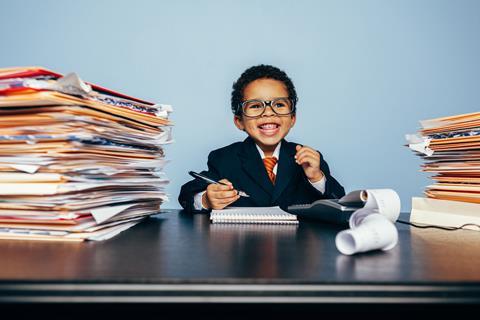 A young boy dressed as a businessman sits at a desk with piles of folders