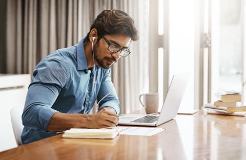 A young man takes notes as he listens to something on a laptop