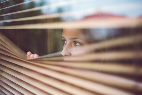 A young woman peers through window blinds