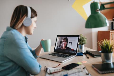 A woman wearing headphones and holding a drink talks to her colleague on a video call